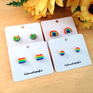 May include: A collection of colorful stud earrings displayed on white cards. The earrings feature designs like rainbows, cat faces, and geometric shapes in various colors. The cards are labeled with the text "hale and hopeful".