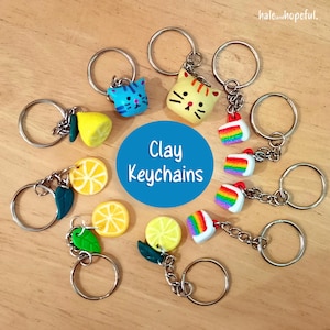 May include: A selection of handmade clay keyrings with diverse designs. Keyrings include a blue cat, a yellow cat, lemon slices, lemon halves, and rainbow cake slices. The keyrings are attached to silver key rings. The text "Clay Keychains" is in a blue circle.