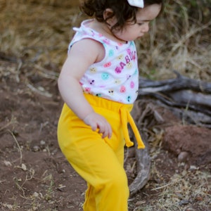Puede incluir: Un niño con una camiseta sin mangas blanca con caras sonrientes de colores y la palabra "BABY" en rosa. También lleva pantalones deportivos amarillos y sandalias marrones. Un lazo blanco adorna su cabello.