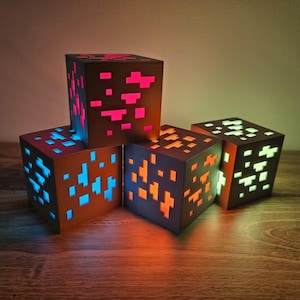 Könnte beinhalten: Fünf verschiedene farbige Minecraft-Block-Nachtlichter. Die Lichter sind aus braunem Material gefertigt und haben ein pixeliges Design. Die Lichter leuchten in verschiedenen Farben, darunter Blau, Grün, Orange, Pink und Weiß.