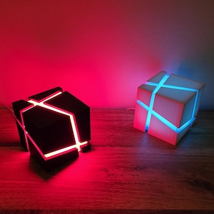 Puede incluir: Dos lámparas geométricas en forma de cubo con luces LED brillantes. La lámpara de la izquierda es negra con luz roja, y la lámpara de la derecha es blanca con luz azul.