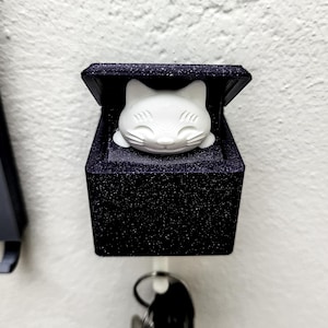 Op de afbeelding: Een zwarte sleutelhanger met een wit kattenhoofd aan de voorkant. De kat heeft een glimlachend gezicht en gesloten ogen. De sleutelhanger heeft een haak aan de onderkant om sleutels aan te hangen.