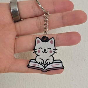 Puede incluir: Un llavero con un gato de dibujos animados leyendo un libro. El gato es blanco con detalles negros y un pequeño sombrero. El libro está abierto con páginas rosas. El llavero está sujeto a una cadena y un anillo plateados.