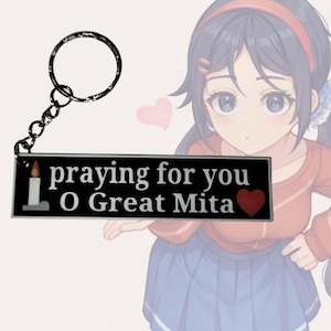 May include: A black and white keychain with the text "praying for you O Great Mita" and a small candle and heart icon. The keychain is attached to a silver key ring. The keychain is held by a cartoon anime girl with blue hair and a red and white striped sweater.