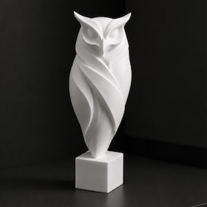 Può includere: Una statuetta di gufo bianco opaco caratterizzata da linee geometriche nette, angolari e fluide che formano il corpo e le ali dell'uccello. Il gufo è appoggiato su una solida base quadrata, presentando una superficie liscia e uniforme.