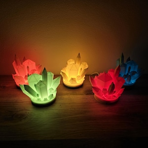 May include: Five crystal-shaped lamps with different colored lights. The lamps are arranged in a row on a wooden surface. The colors of the lights are red, yellow, green, blue, and red.