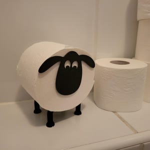 Puede incluir: Un porta rollo de papel higiénico blanco con forma de oveja con ojos negros y nariz negra. El soporte está sobre una encimera blanca con un rollo de papel higiénico al lado.