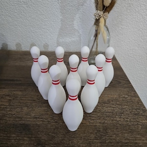 Puede incluir: Un juego de diez bolos blancos con rayas rojas dispuestos en forma de triángulo sobre una superficie de madera.