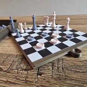 Op de afbeelding: Een gecombineerde schaak- en damspelset. Het bord heeft een zwart-wit geblokt patroon met grijze en roze damstenen. Schaakstukken zijn grijs en wit. De set staat op een houten oppervlak.