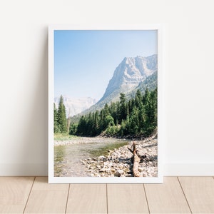 May include: A scenic photograph of a river flowing through a valley with a mountain range in the background. The river is surrounded by trees and rocks. A fallen log lies in the river.