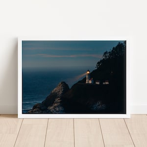 Puede incluir: Un faro blanco con una luz brillante que brilla hacia el mar, encaramado en un acantilado rocoso contra un cielo azul oscuro. El faro está silueteado contra la puesta de sol.