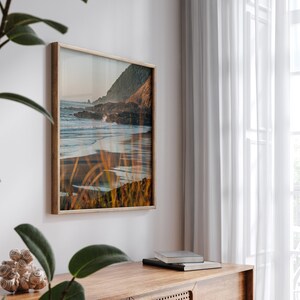 Puede incluir: Una fotograf&iacute;a enmarcada de una escena de playa con una costa rocosa, olas rompiendo en la orilla y hierbas altas en primer plano. La foto est&aacute; en un marco de madera claro y est&aacute; colgada en una pared blanca.