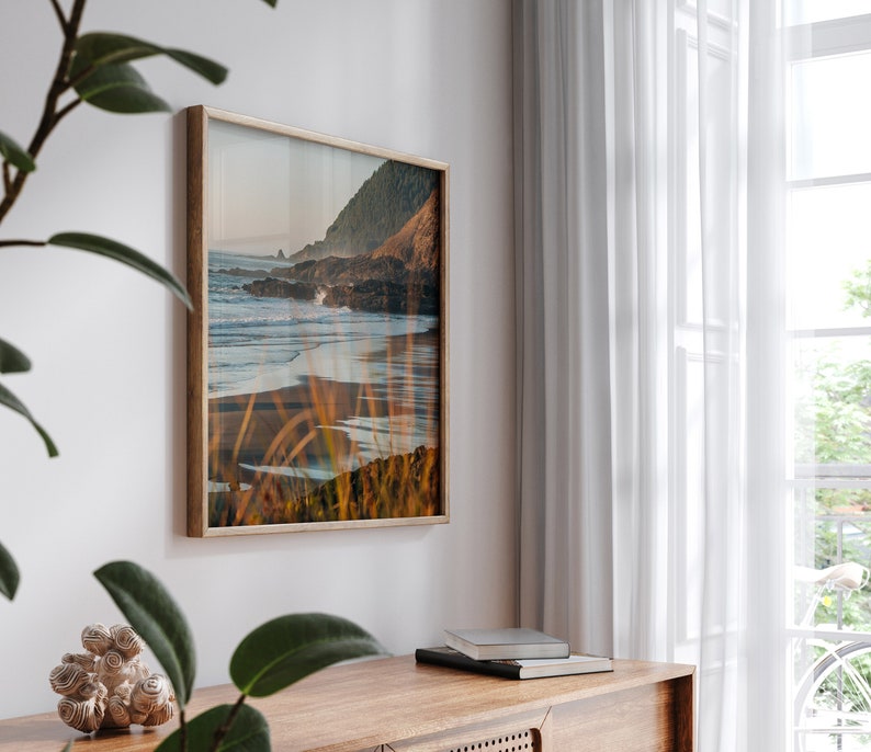 Puede incluir: Una fotograf&iacute;a enmarcada de una escena de playa con una costa rocosa, olas rompiendo en la orilla y hierbas altas en primer plano. La foto est&aacute; en un marco de madera claro y est&aacute; colgada en una pared blanca.