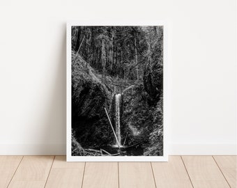 May include: A black and white photograph of a waterfall cascading down a rocky cliff face in a lush forest. The water flows through a narrow opening in the rocks, creating a dramatic scene.