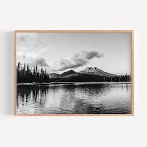 May include: A black and white photograph of a mountain lake with a mountain range in the background. The lake is calm and reflects the sky and mountains. There are trees on the shore of the lake.