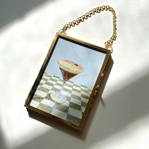 May include: A framed watercolor painting of a cocktail in a martini glass. The painting is set against a checkered background and a light blue sky. The frame is gold-colored, with a delicate gold chain for hanging.