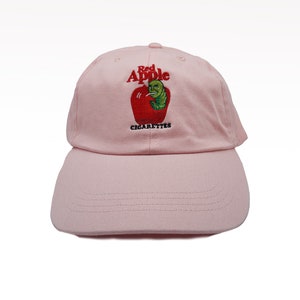 May include: Pink baseball cap with an embroidered red apple and a green worm smoking a cigarette. The text "Red Apple Cigarettes" is embroidered below the apple.