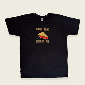 May include: Black t-shirt featuring the text "DAMN GOOD CHERRY PIE" in gold. Below the text is a slice of cherry pie with a golden crust and red filling. A casual, food-themed design.