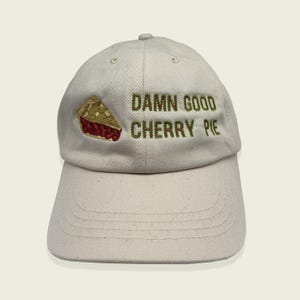 May include: A cream-coloured baseball cap with the embroidered text "DAMN GOOD CHERRY PIE" in green. A slice of cherry pie is embroidered on the left side. The cap has a curved brim and a fabric strap closure.