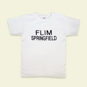 May include: White t-shirt featuring the black text "FLIM SPRINGFIELD" across the front. The shirt has a crew neck and short sleeves. The background is a pale beige colour.