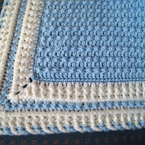May include: A crocheted baby blanket with a blue and white color scheme. The blanket has a textured pattern and a white border.
