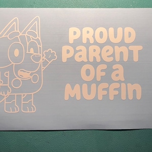 May include: A white vinyl decal featuring a cartoon dog with the text "Proud Parent of a Muffin".