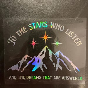Op de afbeelding: Holografische sticker met de tekst "To the stars who listen and the dreams that are answered." Het ontwerp toont een bergketen en drie sterren in roze, oranje en groene tinten. De tekst is in zilver.
