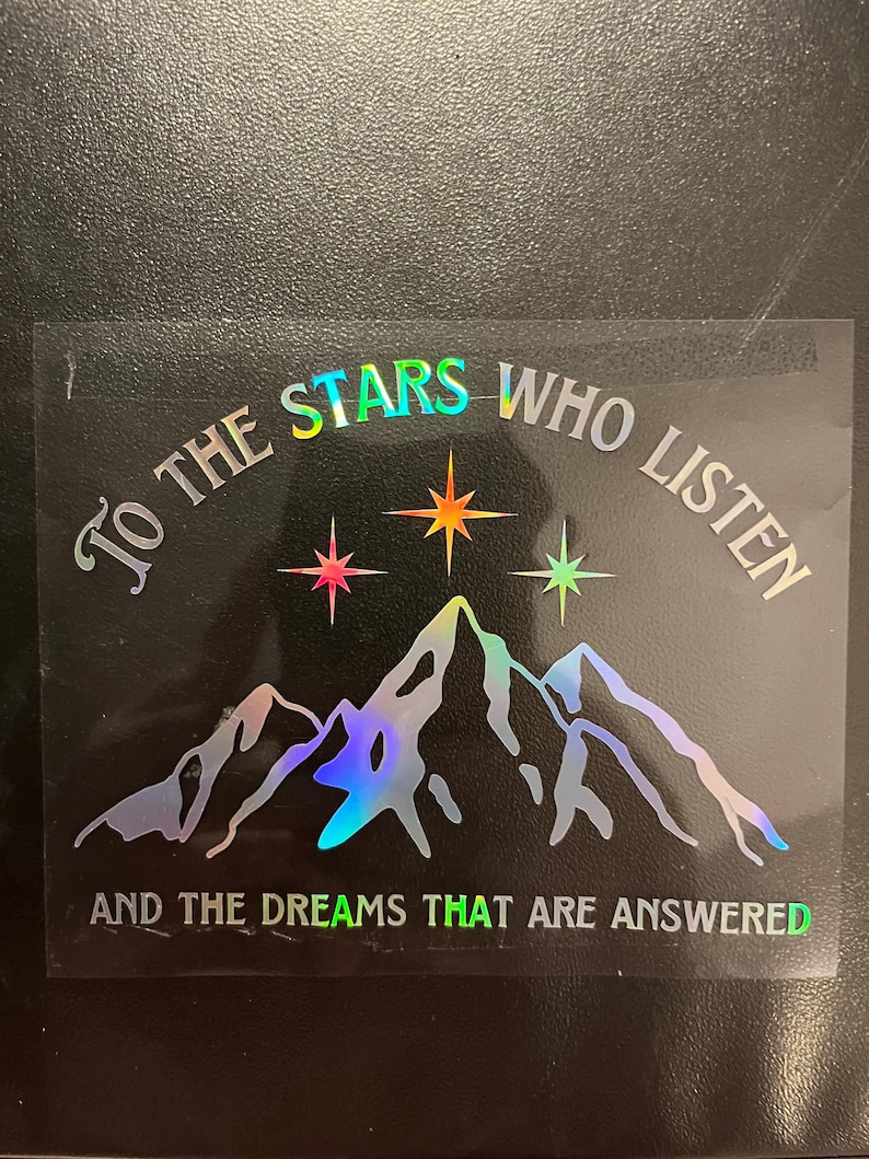 Op de afbeelding: Holografische sticker met de tekst "To the stars who listen and the dreams that are answered." Het ontwerp toont een bergketen en drie sterren in roze, oranje en groene tinten. De tekst is in zilver.
