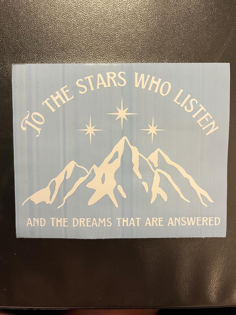 Op de afbeelding: Een lichtblauwe sticker met witte tekst en een bergketenontwerp. De tekst luidt "TO THE STARS WHO LISTEN" boven de bergen en "AND THE DREAMS THAT ARE ANSWERED" eronder.