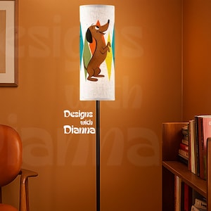 May include: A floor lamp with a cylindrical white shade featuring a brown dachshund illustration and colorful geometric shapes. The lamp has a thin black metal pole and a circular black base, standing upright as a functional lighting fixture.