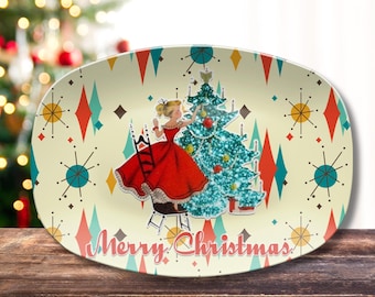 May include: An oval serving platter with a cream background featuring a repeating pattern of red and teal diamonds and starbursts. The center shows a person in a red dress decorating a glittery teal Christmas tree with the text Merry Christmas.