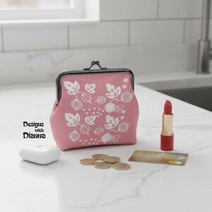May include: A pink coin purse with a metal kiss-lock clasp and a white botanical print featuring gooseberries and leaves. The purse is displayed alongside a white wireless earbud case, a tube of red lipstick, and a credit card on a white surface.