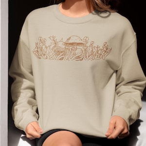 May include: A beige sweatshirt featuring a brown illustration of mushrooms and foliage. The design is centered across the chest. The long-sleeved crew neck top is made of a soft material.