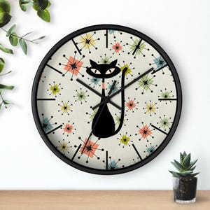 May include: A black-framed wall clock with a mid-century modern design. The clock face features a black cat silhouette and colorful starburst patterns on a cream background. The clock hands are black.