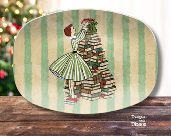 May include: An oval ceramic platter featuring a vintage-style illustration of a person in a green and white striped dress placing a sprig of holly atop a Christmas tree constructed from stacked books, set against a light green striped background.
