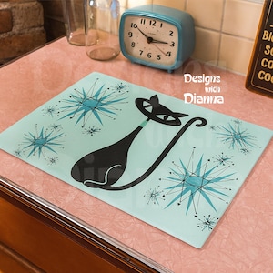May include: A rectangular, light blue cutting board with a black cat design and turquoise starburst patterns. The words "Designs with Dianna" are visible. The board is on a pink surface.