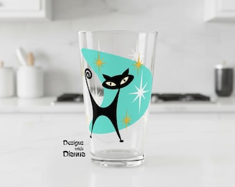 May include: A clear glass tumbler featuring a black cat silhouette centered on a teal boomerang-shaped graphic with gold starburst accents. The text Designs with Dianna is printed in black near the base of the glass, which has a tapered shape.