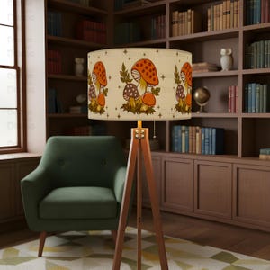 May include: A wooden tripod floor lamp with a beige lampshade featuring a repeating pattern of orange and brown mushrooms. The lamp is in a room with a green armchair and a bookshelf filled with books.