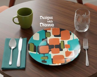 May include: A round plate with a geometric pattern of orange, teal, and olive green rounded squares on a cream background. The setting includes a matte olive green mug, a clear glass, a silver spoon, a silver knife, and a teal napkin.
