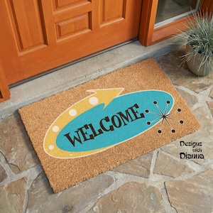 May include: A rectangular tan coir doormat featuring a teal oval shape with a yellow arrow accent. The word WELCOME is printed in black capital letters across the center, accompanied by a black starburst graphic on the right side of the oval.