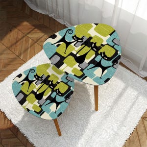 May include: Two mid-century modern style side tables. The tables have a unique, kidney-shaped top with a colorful pattern of black cat silhouettes, green, blue, and gray geometric shapes. The tables have wooden legs and are set on a white rug.
