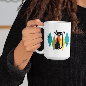 May include: A white ceramic mug featuring a stylized black cat graphic centered among colorful diamond shapes in teal, olive green, and orange, accented by small black starburst patterns on a clean white background surface.