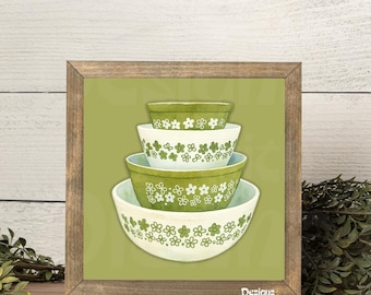 May include: A square framed print featuring a stack of four nesting bowls in varying shades of green and white. Each bowl is decorated with a repeating pattern of small white flowers. The artwork is set against a solid olive green background.