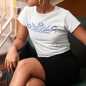 May include: A white t-shirt with a blue floral design on the front. The shirt is being worn by a person sitting on a green chair.