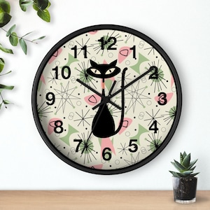 May include: A black and white clock with a black cat design on a cream background with green and pink accents. The clock has a black frame and black hands. The numbers on the clock are black and white.