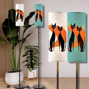 May include: Floor lamps with cylindrical shades featuring a retro design of two stylized black cats against an orange and yellow background. The lamps have black metal stands and round bases. One shade is white, and another is teal.