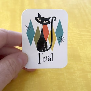 May include: A white rectangular sticker featuring a black cat illustration with a long tail, set against a backdrop of teal, orange, and olive green diamond shapes. The word "Feral" is written in black script below the cat.