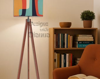 May include: A mid-century modern style floor lamp with a wooden tripod base and a colorful patterned lampshade. The shade features orange, blue, and navy shapes. The text "Designs with Dianna" is visible on the lamp.