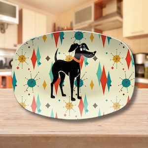 May include: An oval-shaped serving platter featuring a cream-colored background with a repeating pattern of red and teal diamonds, yellow starbursts, and a central illustration of a black dog wearing a simple orange collar facing to the right.