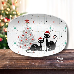 May include: An oval serving platter with a Christmas theme. The platter features a silver Christmas tree with red ornaments, two black cat silhouettes wearing Santa hats, and a geometric pattern. The words "Designs with Dianna" are in the lower right corner.